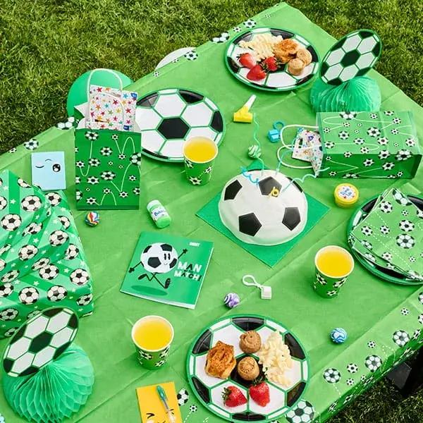 Football-themed party picnic with football-print tableware, party bags, snacks, drinks, and decorations on a green blanket.